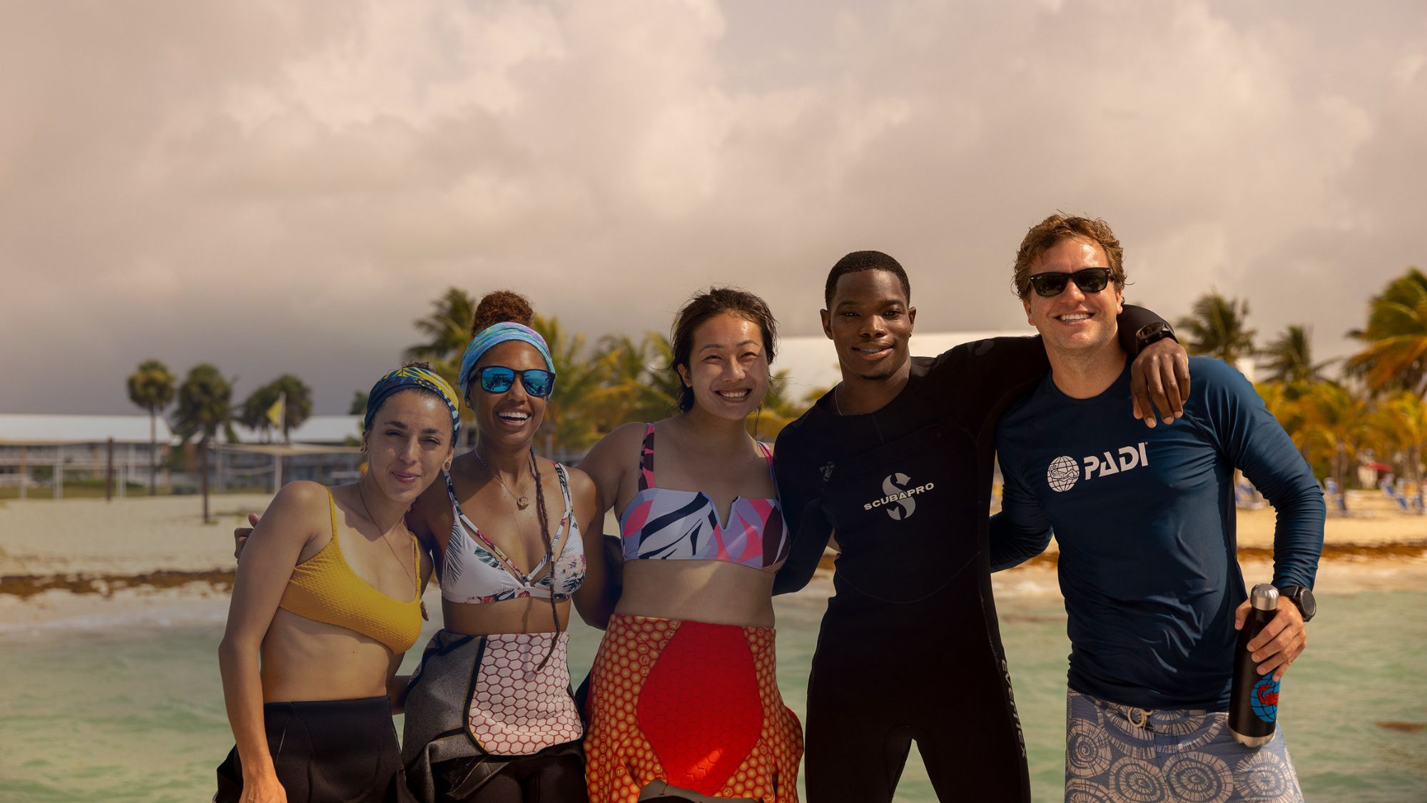 A group of five smiling divers standing together on a beach, wearing wetsuits and swimwear.