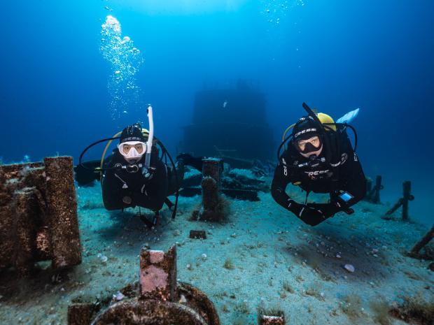 Two divers observe a wreck at the ocean floor.