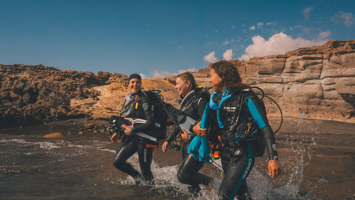 Three women divers walking towards the ocean with scuba gear.